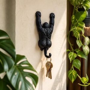 May include: A black, textured wall-mounted hook shaped like a gorilla hanging by its arms. The gorilla's tail curves downwards to form a functional hook, which currently holds a small metal ring with three silver-toned keys attached to it.