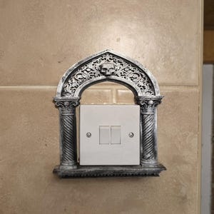 Op de afbeelding: Een decoratieve lichtschakelaar cover met een gotisch boogontwerp. Het zilverkleurige frame heeft een schedel aan de bovenkant en sierlijke details. De cover omringt een witte lichtschakelaar met twee tuimelschakelaars.