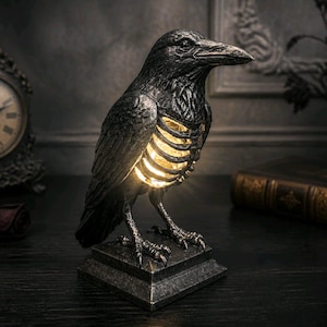 May include: A black raven sculpture with a glowing ribcage, set on a square base. The gothic-style decor includes a vintage clock, a book, and a candlestick. The raven's illuminated ribcage provides a warm glow.