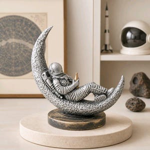 May include: A silver-coloured astronaut figurine resting on a textured crescent moon base. The astronaut wears a detailed space suit and holds a small gold-coloured bottle. The entire sculpture is mounted on a circular, dark, weathered pedestal.