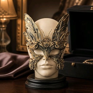 Peut inclure: Un masque décoratif avec des ailes de papillon dorées et noires, présenté sur une tête de mannequin. Le masque présente des motifs complexes et repose sur une base noire, suggérant un thème de mascarade.