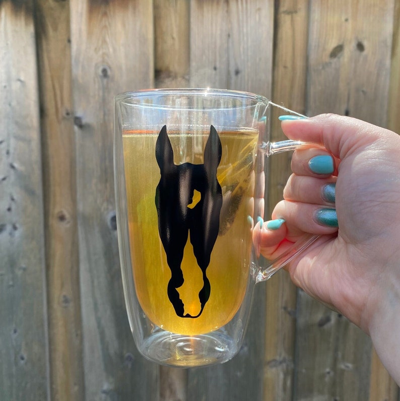Puede incluir: Una taza de vidrio de doble pared con un dise&ntilde;o de silueta de caballo negro. La taza est&aacute; llena de una bebida marr&oacute;n claro.