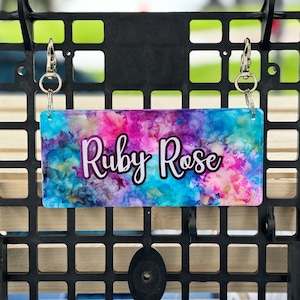 May include: A rectangular keychain with the name "Ruby Rose" in white script, set against a vibrant, abstract background of blues, pinks, and purples. The keychain is attached to a black metal grid with silver clasps.