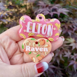 May include: Two dog tags with a peach and banana design. The bone-shaped tag says "ELTON" and the heart-shaped tag says "Raven".