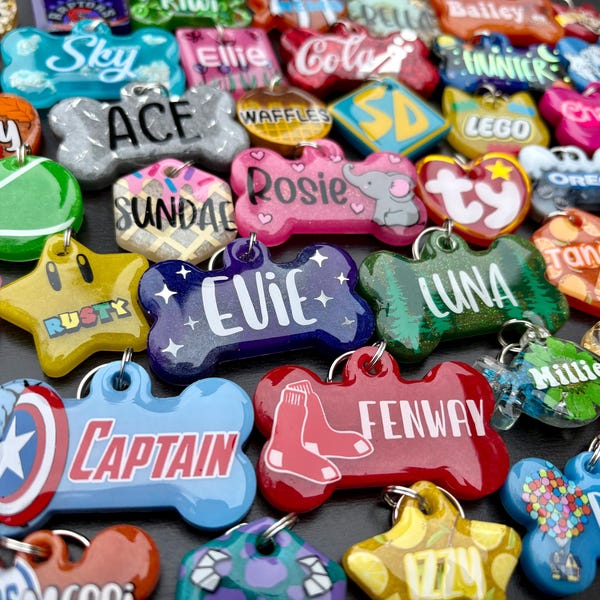 May include: A collection of colorful pet ID tags in various shapes, including bone, star, and heart. The tags feature names like "Evie," "Luna," and "Captain," along with other designs and text. Each tag has a metal ring for attachment.