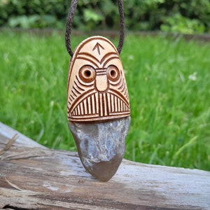 Könnte beinhalten: Ein Holzanhänger mit einem geschnitzten Gesicht und einer grauen Steinfassung. Der Anhänger ist an einer braunen Schnur befestigt und hängt an einer Holzoberfläche.