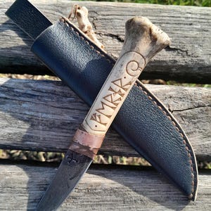 May include: A handmade knife with a bone handle and a leather sheath. The handle is carved with the word "KMK" in runes. The knife has a dark metal blade.