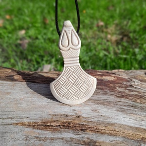 May include: A white wooden pendant in the shape of an axe with a geometric pattern on the blade. The pendant is hanging from a black cord.