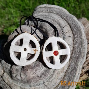 May include: Two white ceramic pendants with a Celtic knot design. One pendant has a red gemstone in the center, and the other has a gold symbol. Both pendants are on a black cord.