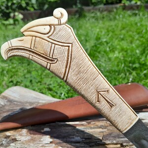 May include: A carved wooden knife handle with an eagle head design. The handle is light brown and has a dark brown leather sheath.
