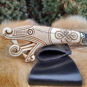 May include: A white bone knife handle with intricate carvings of a dragon and a knot design. The handle is attached to a black leather sheath.