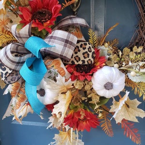 May include: A fall-themed wreath with white pumpkins, sunflowers, and brown and white plaid ribbon. The wreath is decorated with fall leaves and berries.