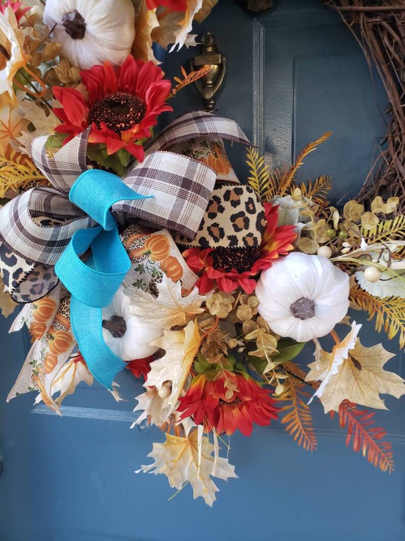 May include: A fall-themed wreath with white pumpkins, sunflowers, and brown and white plaid ribbon. The wreath is decorated with fall leaves and berries.