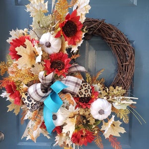 May include: A fall-themed wreath with a brown vine base, decorated with white pumpkins, red sunflowers, brown and leopard print ribbon, and a turquoise bow.