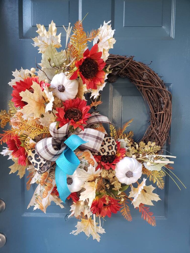 May include: A fall-themed wreath with a brown vine base, decorated with white pumpkins, red sunflowers, brown and leopard print ribbon, and a turquoise bow.