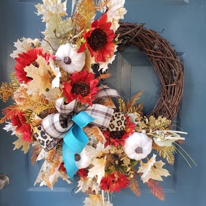 May include: A fall-themed wreath with a brown vine base, decorated with red and orange sunflowers, white pumpkins, fall leaves, and a blue and brown plaid bow.
