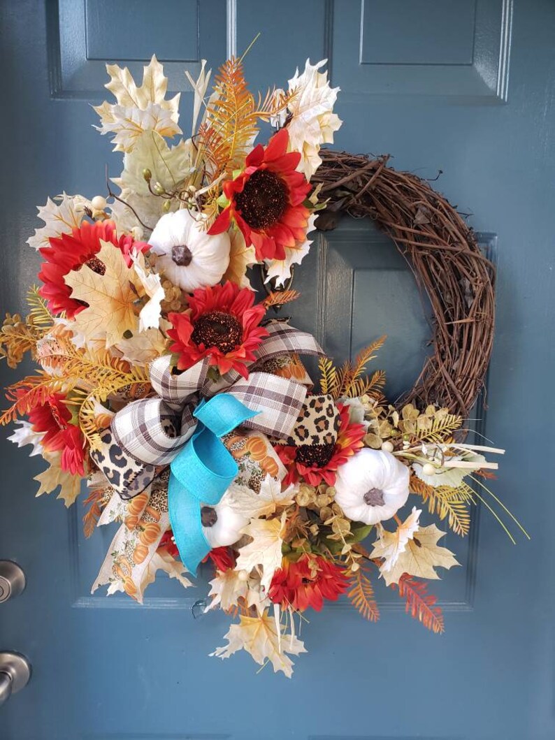 May include: A fall-themed wreath with a brown vine base, decorated with red and orange sunflowers, white pumpkins, fall leaves, and a blue and brown plaid bow.