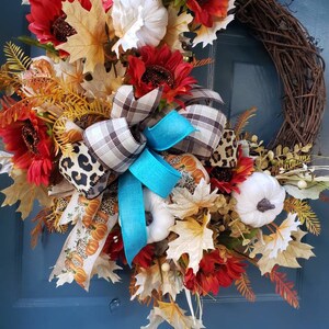 May include: A fall-themed wreath with a brown vine base, decorated with red and orange sunflowers, white pumpkins, and a turquoise and plaid bow.