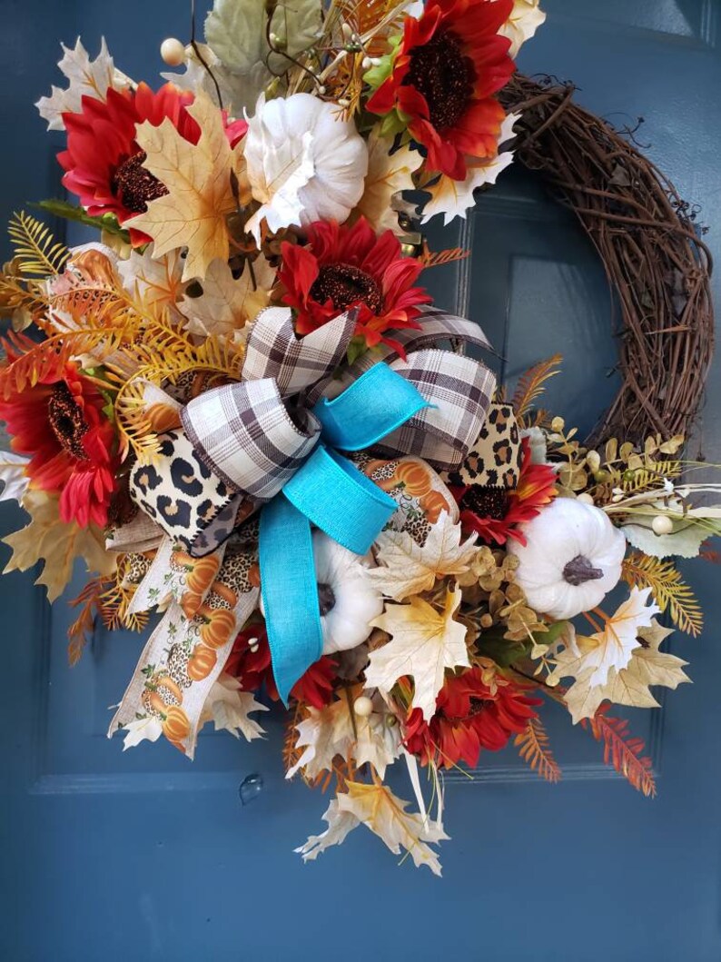 May include: A fall-themed wreath with a brown vine base, decorated with red and orange sunflowers, white pumpkins, and a turquoise and plaid bow.