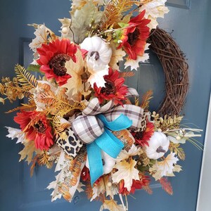 May include: A fall-themed wreath with red sunflowers, white pumpkins, and brown and blue plaid ribbon. The wreath is made of grapevine and has a rustic look.