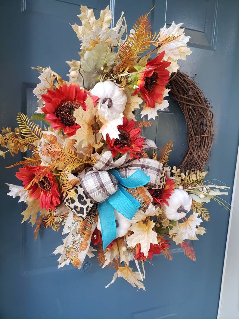 May include: A fall-themed wreath with red sunflowers, white pumpkins, and brown and blue plaid ribbon. The wreath is made of grapevine and has a rustic look.