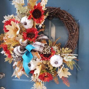 May include: A fall-themed wreath with sunflowers, white pumpkins, and fall leaves. The wreath is decorated with a blue and white plaid ribbon and a leopard print ribbon.