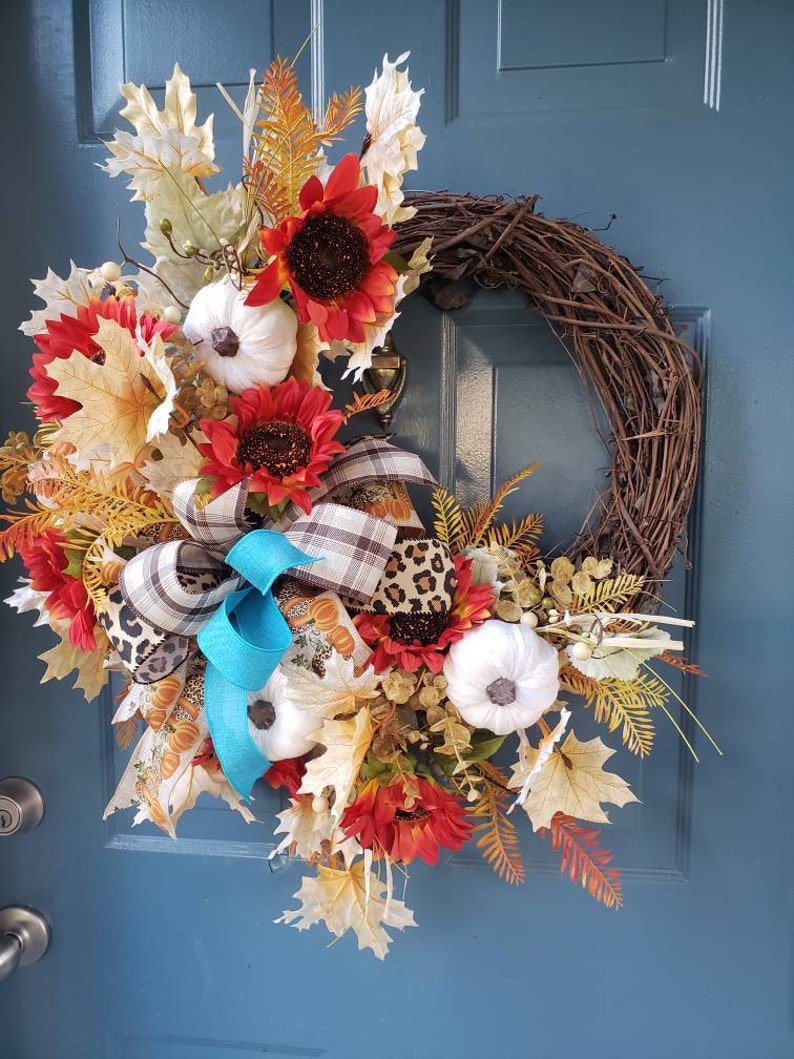 May include: A fall-themed wreath with sunflowers, white pumpkins, and fall leaves. The wreath is decorated with a blue and white plaid ribbon and a leopard print ribbon.