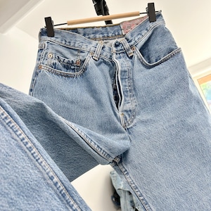 May include: Light blue denim jeans hanging on a wooden hanger. The jeans have a classic five-pocket design, a button fly, and a red and white label on the back pocket. The jeans are a light wash and appear to be vintage.