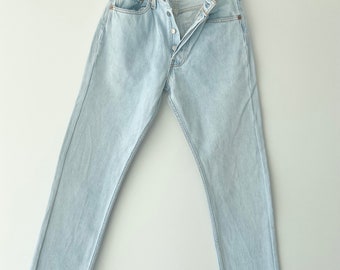 May include: Light blue denim jeans with a button fly and five pockets. The jeans are hanging on a wooden hanger.