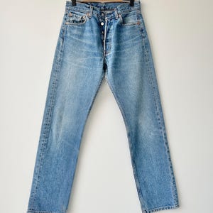 May include: A pair of light blue denim jeans with a button fly and five pockets. The jeans are hanging on a wooden hanger against a white background.