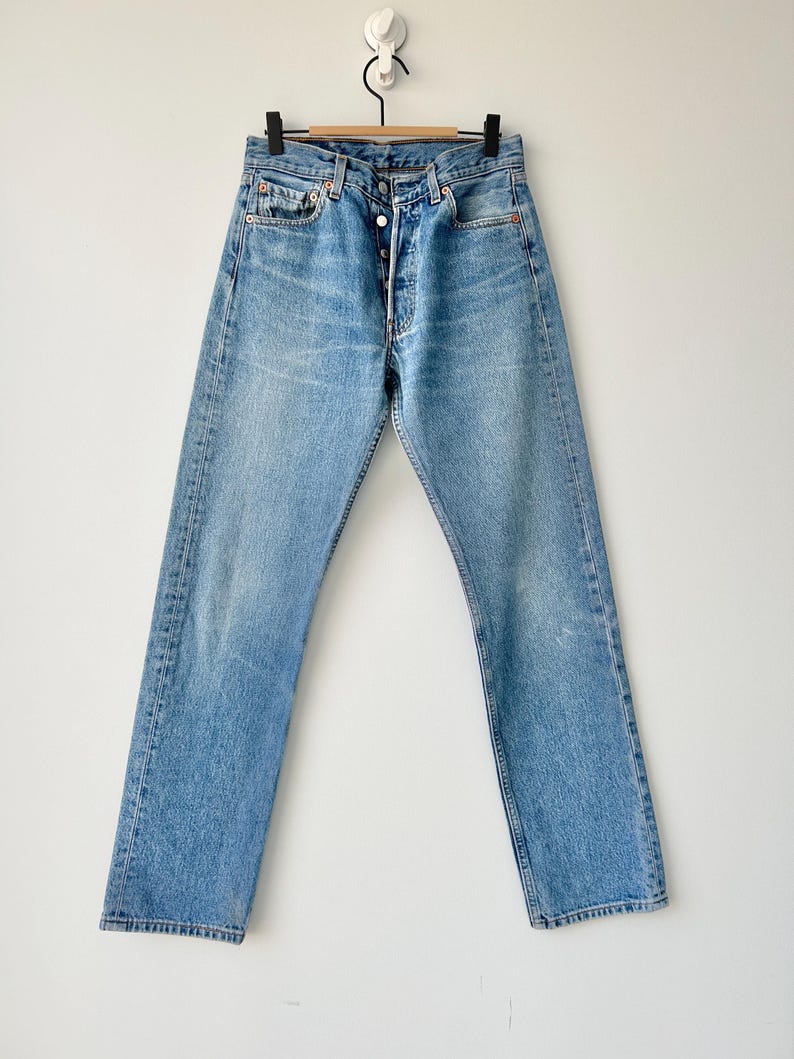 May include: A pair of light blue denim jeans with a button fly and five pockets. The jeans are hanging on a wooden hanger against a white background.