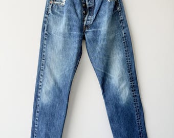 May include: A pair of blue denim jeans featuring a medium wash with intentional fading and distressed edges at the front pockets. The jeans have a button-fly closure, five-pocket styling, belt loops, and copper-colored rivets at the pocket corners.