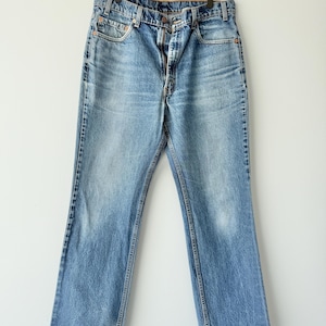 May include: A pair of light blue denim jeans featuring a classic five-pocket design, a button fly closure, and a straight-leg cut. The fabric displays a faded wash with whiskering details at the hips and a worn texture throughout the legs.