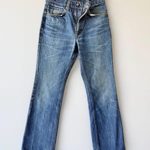 May include: A pair of blue denim jeans with a medium wash, featuring a classic five-pocket design, a button fly closure, and copper-toned rivets. The jeans have a straight-leg cut with subtle whiskering and fading patterns across the front.