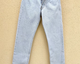 May include: Light blue denim jeans with a button fly and five pockets. The jeans are hanging on a black hanger against a light beige wall.