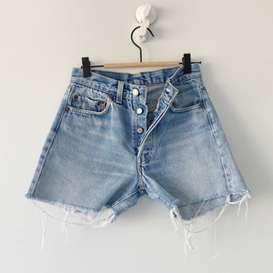 Op de afbeelding: Lichtblauwe denim shorts met een rafelige zoom en een knoopsluiting. De shorts hangen aan een houten hanger.
