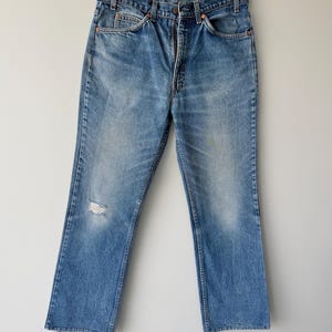May include: A pair of medium-wash blue denim jeans featuring a high-waisted fit, a classic five-pocket design, copper-toned rivets, a button fly closure, and a small distressed rip on the front of the left leg, hanging on a wooden hanger.