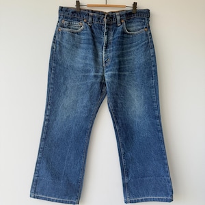 May include: Blue denim jeans with a classic five-pocket design, suspended from a wooden hanger. The jeans feature a button fly and a faded wash, offering a vintage aesthetic. The straight leg cut is visible.