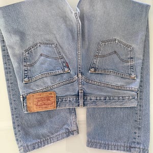 Puede incluir: Vaqueros de mezclilla azul claro con dos bolsillos traseros y una etiqueta de cuero marrón. La etiqueta dice "Levi Strauss & Co." e incluye el número "501". Los vaqueros están doblados cuidadosamente.