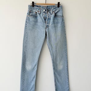May include: Light blue denim jeans with a button fly closure, classic five-pocket design, and a straight leg. The jeans are hanging on a wooden hanger against a white background. The denim has a faded wash.