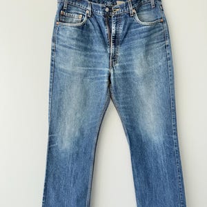 May include: A pair of blue denim jeans featuring a classic five-pocket design, a button fly closure, and copper-toned rivets. The fabric displays a faded wash with whiskering details at the hips and a straight-leg cut, hanging from a hanger.