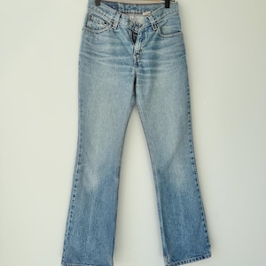 May include: Pale blue denim jeans with a classic five-pocket design and a slight flare at the hem. The jeans are hanging on a wooden hanger against a white wall. The denim has a faded wash.