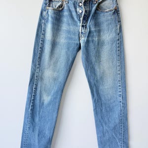 Könnte beinhalten: Hellblaue Jeans mit Knopfleiste, hängen an einem Holzkleiderbügel. Die Jeans weisen Gebrauchsspuren mit verblichenen Bereichen und einem klassischen Fünf-Taschen-Design auf. Der Straight-Leg-Stil ist sichtbar.