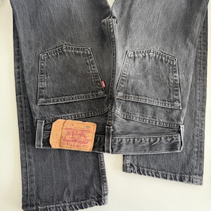 Puede incluir: Dos pares de vaqueros de mezclilla negros, doblados y colocados uno al lado del otro. Los vaqueros tienen un estilo clásico de cinco bolsillos, con una etiqueta roja en el bolsillo trasero. La etiqueta de la marca Levi's es visible en un par.