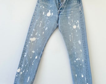 May include: Light blue denim jeans with white paint splatters. The jeans have a button fly and are hanging on a hanger.