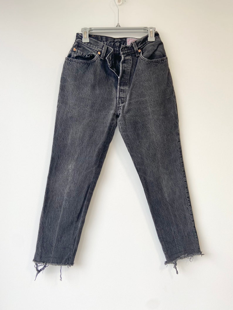 May include: Black denim jeans with a frayed hem. The jeans have a classic five-pocket style and a button fly.