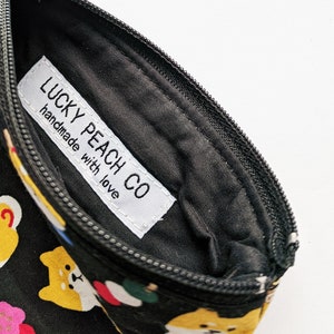 May include: A black zippered pouch with a white label that reads "LUCKY PEACH CO handmade with love". The pouch is decorated with a repeating pattern of yellow Shiba Inu dogs and other Japanese-inspired designs.