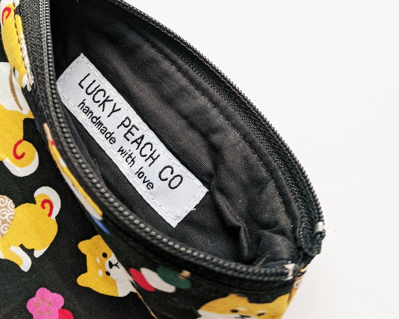 May include: A black zippered pouch with a white label that reads "LUCKY PEACH CO handmade with love". The pouch is decorated with a repeating pattern of yellow Shiba Inu dogs and other Japanese-inspired designs.