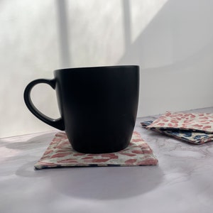 May include: A black ceramic mug with a handle sits on a pink and white patterned coaster. The coaster is on a white marble surface.
