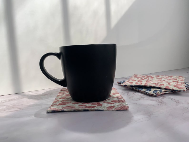 May include: A black ceramic mug with a handle sits on a pink and white patterned coaster. The coaster is on a white marble surface.